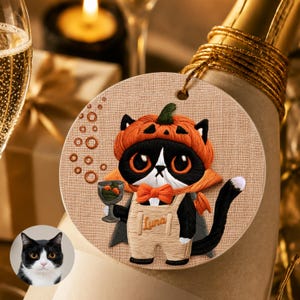 May include: A round beige ornament featuring an embroidered black and white cat wearing an orange pumpkin hat, a matching bow tie, and tan overalls with the name Luna stitched on the front. The cat holds a small glass with orange contents.