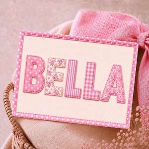 May include: A rectangular fabric sign with a pink polka dot border featuring the name BELLA in large, appliqued letters. The letters B, E, L, L, and A are crafted from various pink fabrics, including polka dots, floral prints, stripes, and gingham patterns.