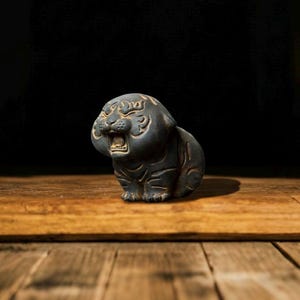 May include: A small, dark grey ceramic tiger figurine with an open mouth, sitting on a wooden surface. The tiger has etched details and a stylized design. The background is a dark, solid colour.