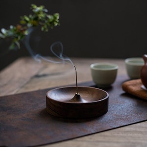 May include: A dark brown wooden incense holder with a burning incense stick, emitting a wisp of smoke. The holder is round with a central cone. The background includes tea cups and a sprig of greenery.