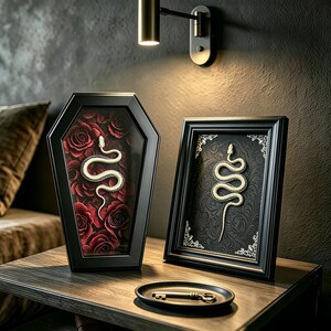 May include: A black coffin-shaped frame containing a white snake on a red rose background, next to a rectangular black frame with a white snake on a black patterned background, and a small black dish holding a single metal key.