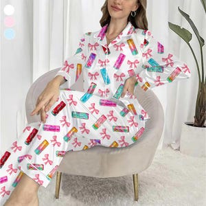 May include: White satin pajama set with a lipstick and pink bow pattern. The long-sleeved top and pants feature a button-down shirt design with white piping. The lipstick designs are in various colors.