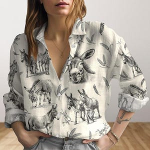 Puede incluir: Camisa abotonada crema con un estampado repetido de ilustraciones de burros en blanco y negro. La camisa de manga larga también presenta espigas de trigo. Las mangas están dobladas, revelando un puño estampado.