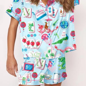 May include: Light blue pajama set with a Mahjong-themed print. The short-sleeved top and shorts feature illustrations of Mahjong tiles, dragons, lanterns, and the text "MAHJONG". The set has pink trim and buttons.
