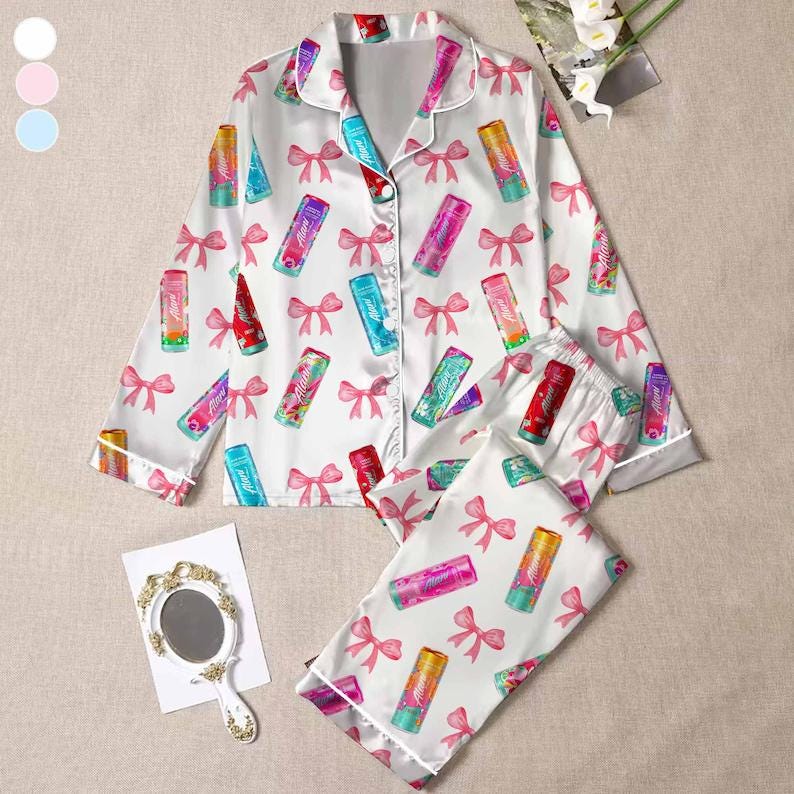 May include: White pajama set with a lipstick and bow pattern. The long-sleeved top and pants feature colorful lipstick tubes and pink bows. The set has white trim and a button-down front.