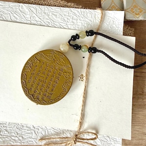 May include: A gold-coloured pendant with Chinese characters and a black cord. The pendant is round with an embossed design and text. The cord has black and green beads. The pendant is on a white card tied with twine.
