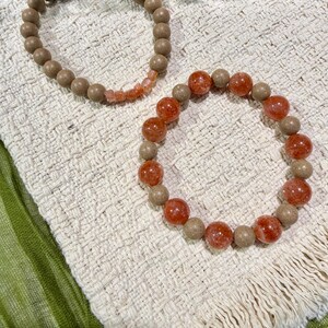 May include: Two beaded bracelets. One is made of light brown, round beads with a section of smaller, orange-pink beads. The other bracelet features a mix of light brown and orange-red beads, creating a contrasting colour scheme.