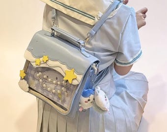 Könnte beinhalten: Hellblaue Umhängetasche im Satchel-Stil mit Wolken- und Sterndesign. Die Tasche hat eine transparente Frontplatte mit einer Cartoonfigur und Perlenakzenten. Gelbe Sterne und weiße Wolken verzieren die Oberseite. Enthält einen kleinen Plüschanhänger.