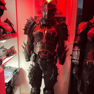 May include: A black and red Batman costume with a helmet, chest armour, and spiked accents. The costume has a bat symbol on the chest and is displayed in a room with other Batman-themed items.