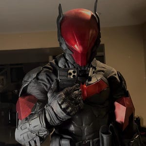 Op de afbeelding: Een tactisch pak voor het hele lichaam met een gladde, glanzende rode helm met puntige oren, een zwarte borstplaat met een rood embleem, gesegmenteerde zwarte bepantsering op de armen en schouders, zwarte tactische handschoenen en een gereedschapsriem met holsters.