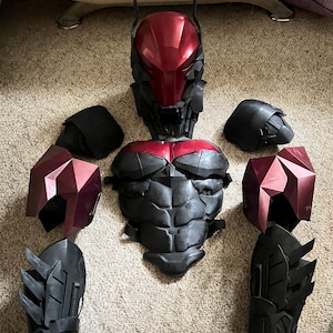 May include: A disassembled black and red superhero costume, including a helmet, chest piece, shoulder pads, and gauntlets. The helmet and shoulder pads have a faceted design, while the chest piece has a sculpted appearance. The costume is arranged on a neutral-colored carpet.