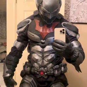Könnte beinhalten: Ein Batman Beyond-Ganzkörperkostüm mit einem silber-schwarzen Helm mit spitzen Ohren, einem schwarzen Anzug mit silbernen Panzerplatten an Brust, Schultern und Armen, einem roten Fledermaus-Emblem auf der Brust und einem Gürtel mit roter Schnalle.