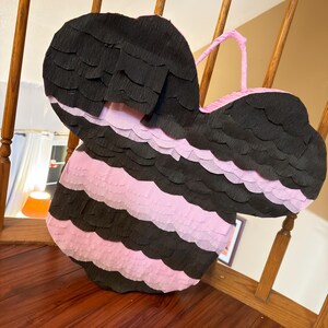 May include: A large, handmade pi&ntilde;ata in the shape of a bee. The pi&ntilde;ata is black and pink, with alternating horizontal stripes. It has a pink handle and is made of crepe paper.