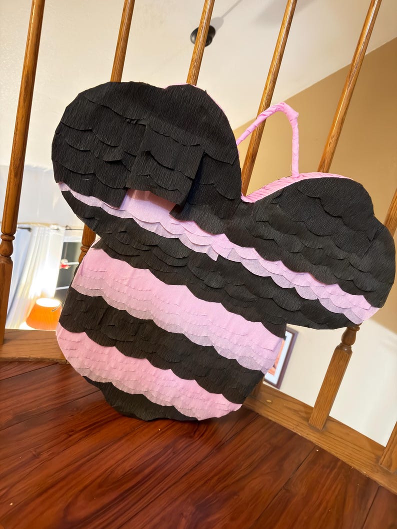 May include: A large, handmade pi&ntilde;ata in the shape of a bee. The pi&ntilde;ata is black and pink, with alternating horizontal stripes. It has a pink handle and is made of crepe paper.