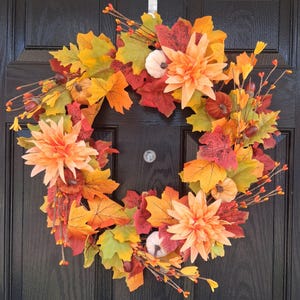 May include: A circular autumn wreath featuring a mix of orange, yellow, and red maple leaves, three light orange dahlia flowers, small white pumpkins, and brown acorns. The wreath is constructed with thin brown stems and small orange berries.