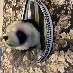 Puede incluir: Una cabeza de panda de peluche caprichosa con un diseño de payaso. El panda tiene marcas negras y moradas en los ojos, acentos verdes y una nariz negra. Está adornado con un sombrero a rayas, un borde de cuentas y un cuello con volantes, creando una pieza decorativa única.