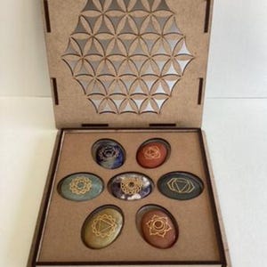Könnte beinhalten: Eine Holzbox mit einem geometrischen Design auf dem Deckel, geöffnet, um sieben ovale Chakra-Kristalle zu enthüllen. Jeder Kristall hat eine andere Farbe und ein anderes Symbol. Der Text unten lautet "7 Chakra Crystal Set, Balance, Energy, Harmony."