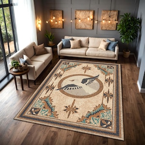 May include: A rectangular area rug with a mosaic design featuring a central circular emblem with stylized wings. The rug has a beige background with brown, teal, and navy blue accents. The rug is placed in a living room setting with beige sofas and wooden floors.