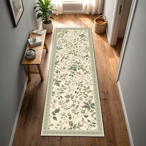 May include: A long rectangular runner rug featuring a cream-colored background with a repeating botanical pattern of sage green leaves and thin branches. The rug is framed by a decorative border with a matching green and cream geometric design.