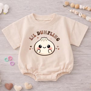 May include: A beige baby romper with the text "Lil Dumpling" above a cartoon dumpling illustration. The romper is surrounded by baby accessories, including a stuffed bunny, shoes, and wooden toys.