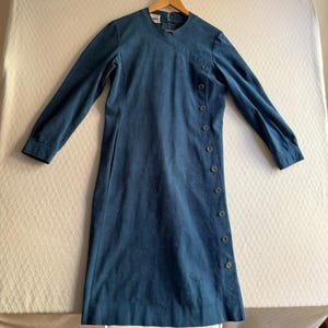 May include: A long-sleeved, knee-length dress in a solid teal blue color. The garment features a high neckline with an asymmetrical wrap-style front closure secured by a vertical row of nine matching buttons extending down the left side.