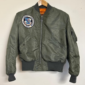 May include: Olive green bomber jacket with a full zipper closure, ribbed cuffs, and a collar. Features a patch on the left chest with text and a design. The jacket has two front pockets and a small pocket on the left sleeve. The interior lining is orange.