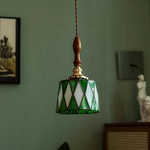 May include: A hanging pendant light with a stained glass shade. The shade features a green and white diamond pattern. The light has a wooden top and a brass base. The cord is brown and twisted.