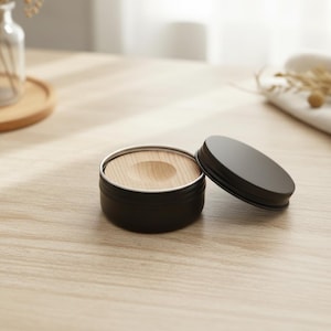 May include: A black metal container with a silver rim and a wooden insert. The lid is off to the side. The container sits on a light-colored wooden surface. The container is round and appears to be a small storage tin.
