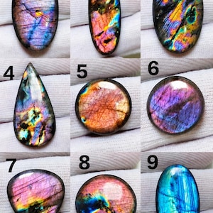 Pode incluir: Uma coleção de nove pedras preciosas de labradorite polidas numeradas de um a nove. Cada pedra apresenta uma superfície iridescente única com flashes vibrantes de azul, roxo, laranja e amarelo, exibindo fraturas e padrões naturais.