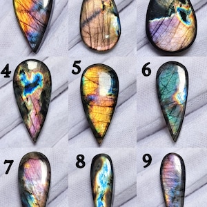 May include: Nine teardrop-shaped labradorite gemstones with iridescent colors. Each stone displays a unique pattern of blues, greens, yellows, and purples. The polished stones are displayed against a white background, showcasing their natural beauty.