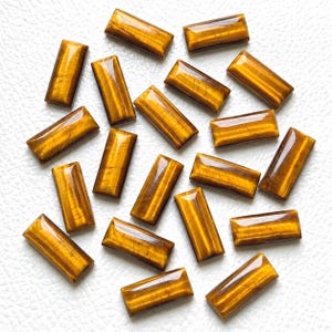 May include: A collection of seventeen rectangular tiger's eye gemstones, each featuring a polished surface with a characteristic chatoyant golden-brown banded pattern. These smooth, flat-backed cabochons are arranged in a scattered formation.