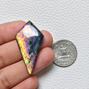 May include: A kite-shaped labradorite gemstone featuring iridescent flashes of yellow, orange, and blue, displayed next to a United States quarter dollar coin for scale, showcasing its polished surface and natural mineral inclusions.