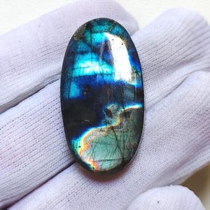 May include: An oval-shaped labradorite gemstone with a polished surface. The stone displays a vibrant play of color, with iridescent flashes of blue, teal, and hints of yellow and orange. The gemstone is set against a white background.
