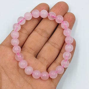 May include: A rose quartz beaded bracelet. The bracelet features round, light pink beads strung together on a pink elastic cord. The beads are smooth and polished, reflecting light. The bracelet is shown on a hand.