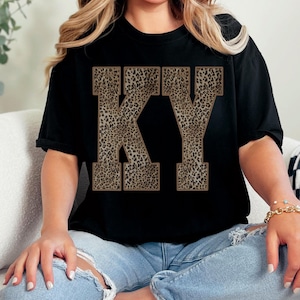May include: A black short-sleeved t-shirt featuring the letters KY in a large, block-style font. The letters are filled with a brown and black leopard print pattern. The shirt is paired with light blue denim jeans that have distressed knee areas.