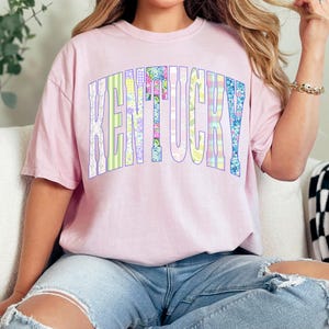 May include: A light pink t-shirt with the word "KENTUCKY" in large, colorful, patchwork-style letters. The letters are outlined in a light purple. The shirt is paired with blue jeans.