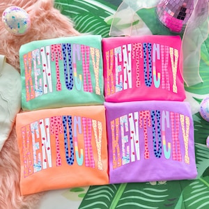 May include: Four colourful sweatshirts are stacked, each with "Kentucky" in a decorative, arched font. The shirts are in shades of mint green, pink, orange, and purple. The letters have various patterns, including stars and polka dots.