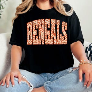 Puede incluir: Una camiseta negra de manga corta con la palabra BENGALS impresa en letras grandes y arqueadas con un patrón de rayas de tigre naranja y blanco en el pecho. La prenda tiene cuello redondo y un corte holgado, combinada con vaqueros azul claro desgastados.