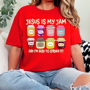 May include: Red t-shirt with the text "JESUS IS MY JAM" and illustrations of jam jars labeled "Faith", "Pray", "Blessed", "Eternal", "Love", "Gratitude & Grace", "Hope", and "Believe". The text "AND I'M HERE TO SPREAD IT!" is also included.
