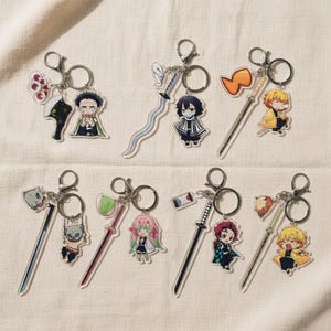 Könnte beinhalten: Sammlung von Anime-Schlüsselanhängern. Jeder Schlüsselanhänger zeigt einen Charakter oder ein Objekt aus der Serie, darunter Schwerter, Figuren und anderes Zubehör. Die Schlüsselanhänger sind aus klarem Acryl gefertigt und haben silberfarbene Metall-Schlüsselringe.