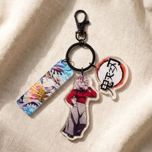 Puede incluir: Un llavero con tres colgantes de estilo anime. Un colgante tiene forma rectangular con un personaje y un diseño de abanico. Otro colgante es un personaje con un atuendo rojo. El tercer colgante es un bocadillo con texto japonés.