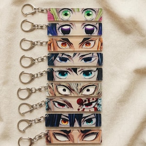 Könnte beinhalten: Eine Sammlung rechteckiger Schlüsselanhänger mit Anime-Augen-Designs. Jeder Schlüsselanhänger zeigt ein einzigartiges Augenpaar mit unterschiedlichen Farben und Stilen, befestigt an einem silbernen Schlüsselring. Die Schlüsselanhänger sind auf einem neutralfarbenen Stoff angeordnet.
