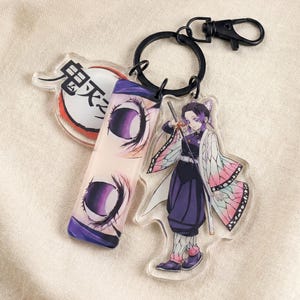 Può includere: Un portachiavi in acrilico trasparente con illustrazioni in stile anime. Il portachiavi include un personaggio con i capelli viola e una spada, un ciondolo rettangolare con occhi viola e un ciondolo circolare con testo giapponese. È attaccato a un anello e un fermaglio in metallo nero.