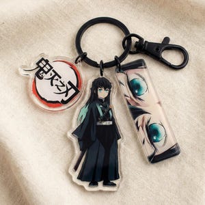 Könnte beinhalten: Ein schwarzer Schlüsselanhänger mit drei Acryl-Anhängern. Ein Anhänger zeigt eine Figur in schwarzer Kleidung mit türkisfarbenem Haar, ein anderer hat ein kreisförmiges Design mit japanischem Text, und der letzte zeigt eine Nahaufnahme von Augen. Der Schlüsselanhänger hat einen schwarzen Ring und Verschluss.