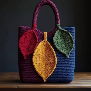 May include: A crochet tote bag featuring a deep blue and burgundy body with a matching burgundy handle. The front is decorated with three large, textured, leaf-shaped appliques in vibrant red, mustard yellow, and forest green colors.