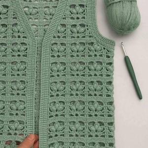 May include: A sage green crochet vest with a floral pattern. The sleeveless garment has a V-neck and a buttonless front. A ball of matching yarn and a crochet hook are next to the vest.