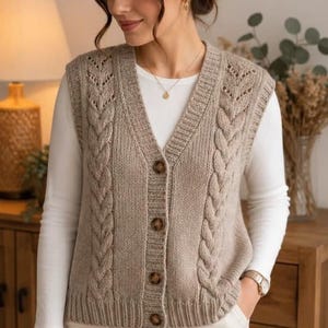May include: A beige knit sweater vest featuring a V-neckline, a front button closure with four brown buttons, and vertical cable knit patterns along the front panels. The garment is worn over a plain white long-sleeved shirt and white pants.
