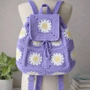 May include: A crocheted backpack in a light purple hue, adorned with white and yellow daisy motifs. The bag features adjustable straps, a drawstring closure, and a flap. The handmade bag is displayed on a mannequin.