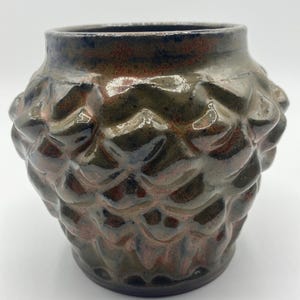 May include: A handcrafted ceramic vase with a textured, scale-like pattern. The vase features a dark, earthy color palette with shades of brown, green, and hints of blue. The opening is a dark blue, and the vase has a rounded shape.