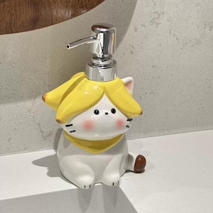 Könnte beinhalten: Ein Keramik-Seifenspender in Form einer weißen Katze mit gelbem Bananenschalenhut und Schal. Der Spender hat eine silberne Pumpe und schwarze Augen. Die Katze sitzt auf einer weißen Oberfläche.